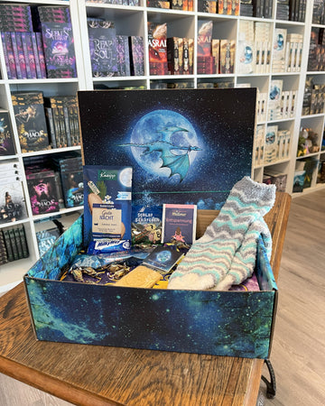 Das Produktbild zeigt vorn eine geöffnete Drachenpost in blauem Schuppen-Design. Man sieht im Deckel der Box das Verlagslogo - einen Drachen, der vor dem Mond fliegt - und in der Box die zum Thema Drachentraum passenden Goodies: Kuschelsocken, Badezusatz, Gute-Nacht-Tee und andere Kleinigkeiten. Im Hintergrund sieht man die Bücherregale des Verlagsladens.